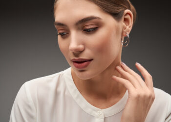 A young woman in a white blouse, touching her neck, with a silver earring.